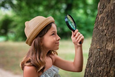 Young girl in a sundress and straw hat holding up a magnifying glass to examine a tree trunk, illustrating that Christina Winkler offers online therapy for children ages 7-12.